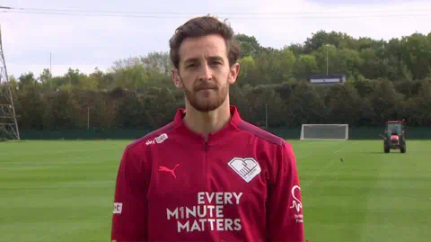 BBC Tom Lockyer, with brown hair and beard, is standing on a football pitch wearing a red football top which says "Every Minute Matters"